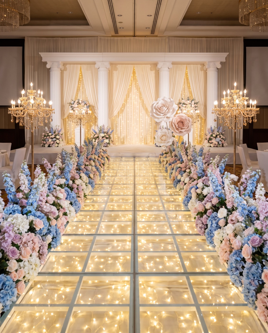 Glass light walkway lined with blue and blush silk flower hedges leading to a pillar stage backdrop
