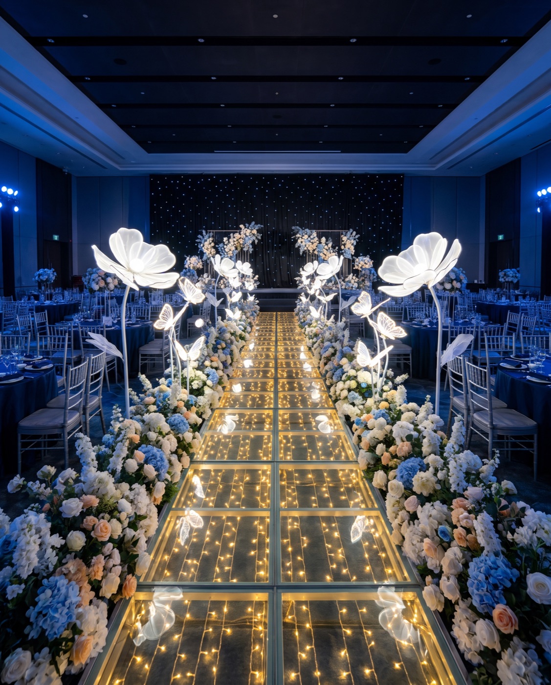 Giant glowing flower lamps and blue flower hedges along a lit glass walkway