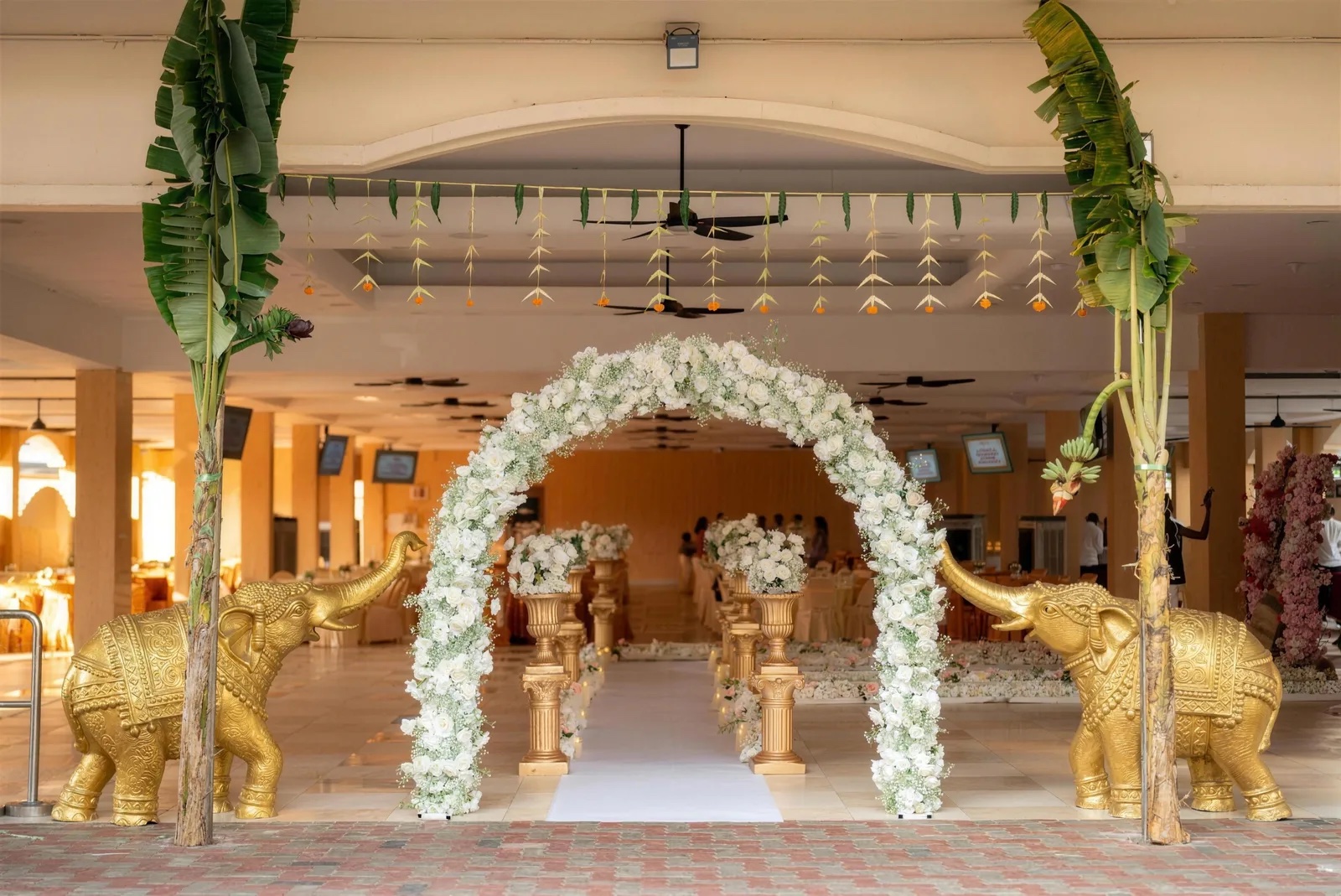 White floral entrance arch with gold elephants and banana trees at PGP Hall