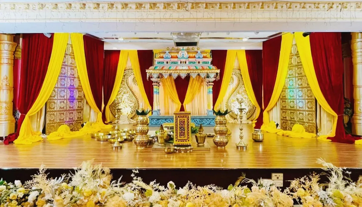 A temple-style wedding stage with red and yellow drapes and brass kalasam, by Alangkaar